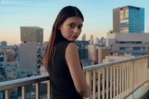 Young woman in black top posing on JG Studio balcony with cityscape background.