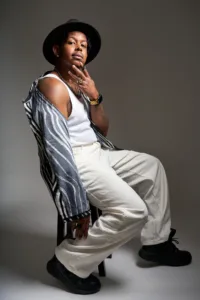 Man in a black fedora, white tank top and patterned shirt off one shoulder, posing on a studio stool.