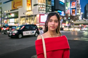 Portrait photo session in Shibuya at night, woman in red off-shoulder sweater with Tokyo neon lights and traffic in the background: JG Studio Tokyo photography tour.